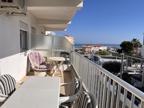 Ein Balkon mit Tisch und Stühlen, ein Fenster mit Jalousien und Blick auf das Meer und die Gebäude.