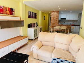 Ein Wohnzimmer mit einem beigen Sofa, einem Fernseher, einer Anrichte und einer Küchenzeile mit einer Steinsäule.