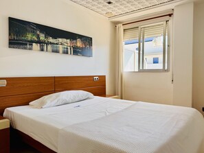 Habitación de hotel con cama, mesita de noche, ventana con persianas y una pintura de un paisaje urbano en la pared.