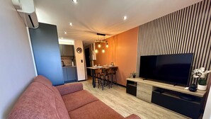 A modern living room with a sofa, a TV, a dining area, and a kitchen in the background. - Apartment port view, air conditioning - 4 guests (Cap d'Agde)