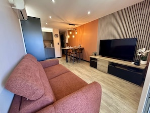 Ein modernes Wohnzimmer mit Sofa, Fernseher, Essbereich und einer Küche im Hintergrund.