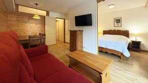 Una habitació amb sòl de fusta, un sofà vermell, una taula de fusta, un llit amb edredó blanc, una televisió penjada a la paret i una porta que dona a una altra habitació.