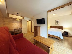 A room with a wooden floor, a red sofa, a wooden table, a bed with a white bedspread, a television mounted on the wall, and a door leading to another room. - Comfortable apartment - 6 guests (Les Orres)
