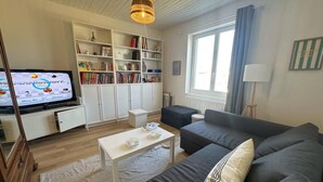 Ein Wohnzimmer mit einer Couch, einem Couchtisch, einem Fernseher, der ein Spiel zeigt, einem Bücherregal und einem Fenster mit Vorhängen.