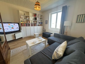 Ein Wohnzimmer mit einer Couch, einem Couchtisch, einem Fernseher, der ein Spiel zeigt, einem Bücherregal und einem Fenster mit Vorhängen.