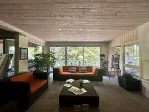 Un vestíbulo amplio con techo de madera, un ventanal con vistas a árboles, una zona de estar con sofá y sillones, y una mesa de centro con revistas.