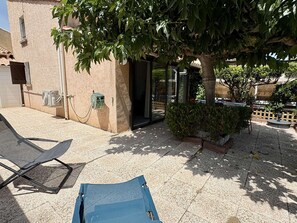 Ein Gebäude mit einer gefliesten Terrasse, einem Baum, der Schatten spendet, und einem Stuhl.