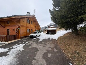 En træbygning med en bil foran, sne på jorden og et træ til højre.