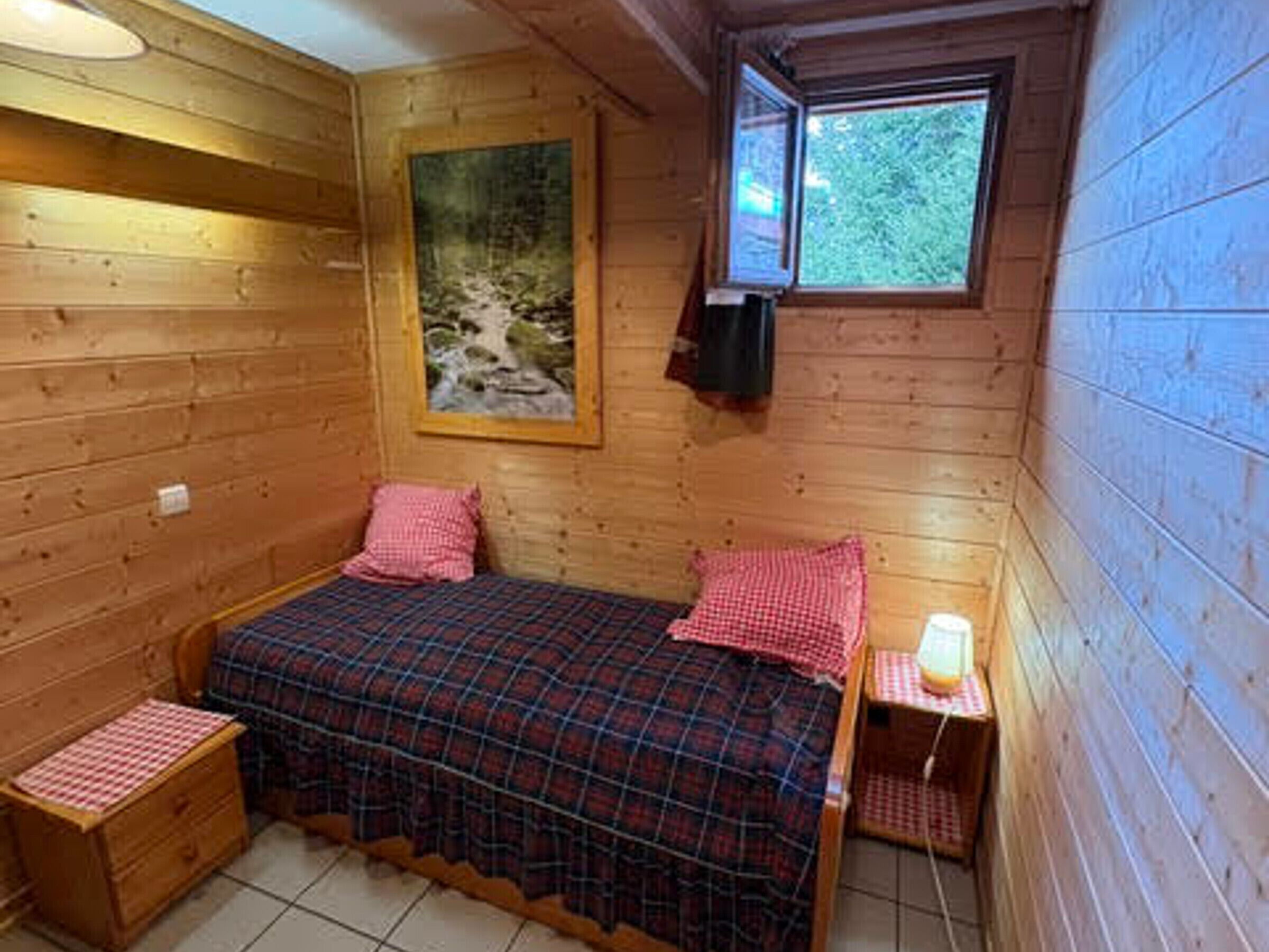 Una pequeña habitación de cabaña de madera con una cama, una mesita de noche, una ventana y un cuadro en la pared.