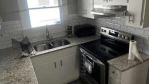 Une cuisine avec des meubles blancs, un fourneau noir, un four à micro-ondes et une fenêtre avec des stores.
