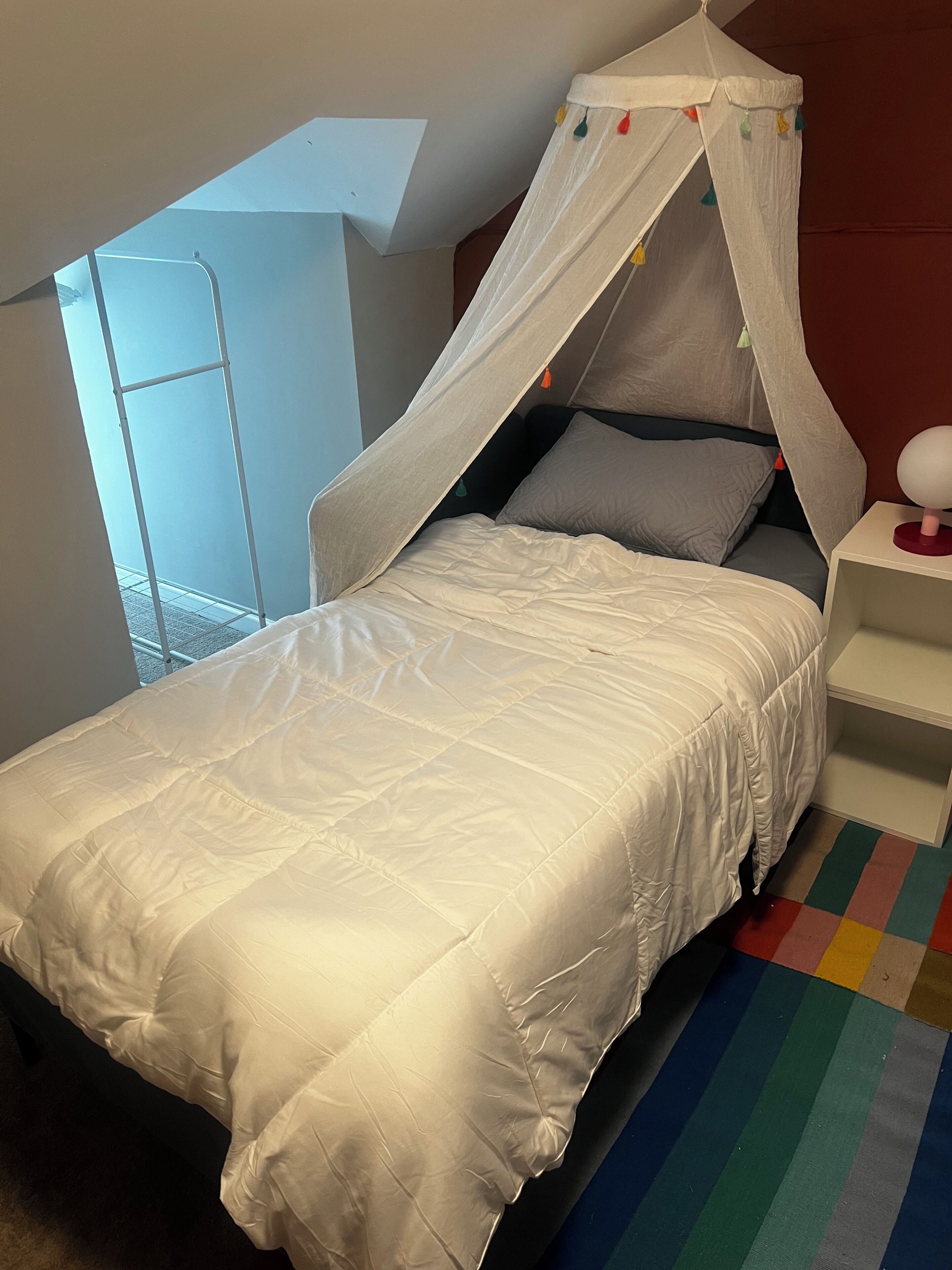 Una cama individual con dosel, una mesita de noche con lámpara y un colorido tapete a rayas.