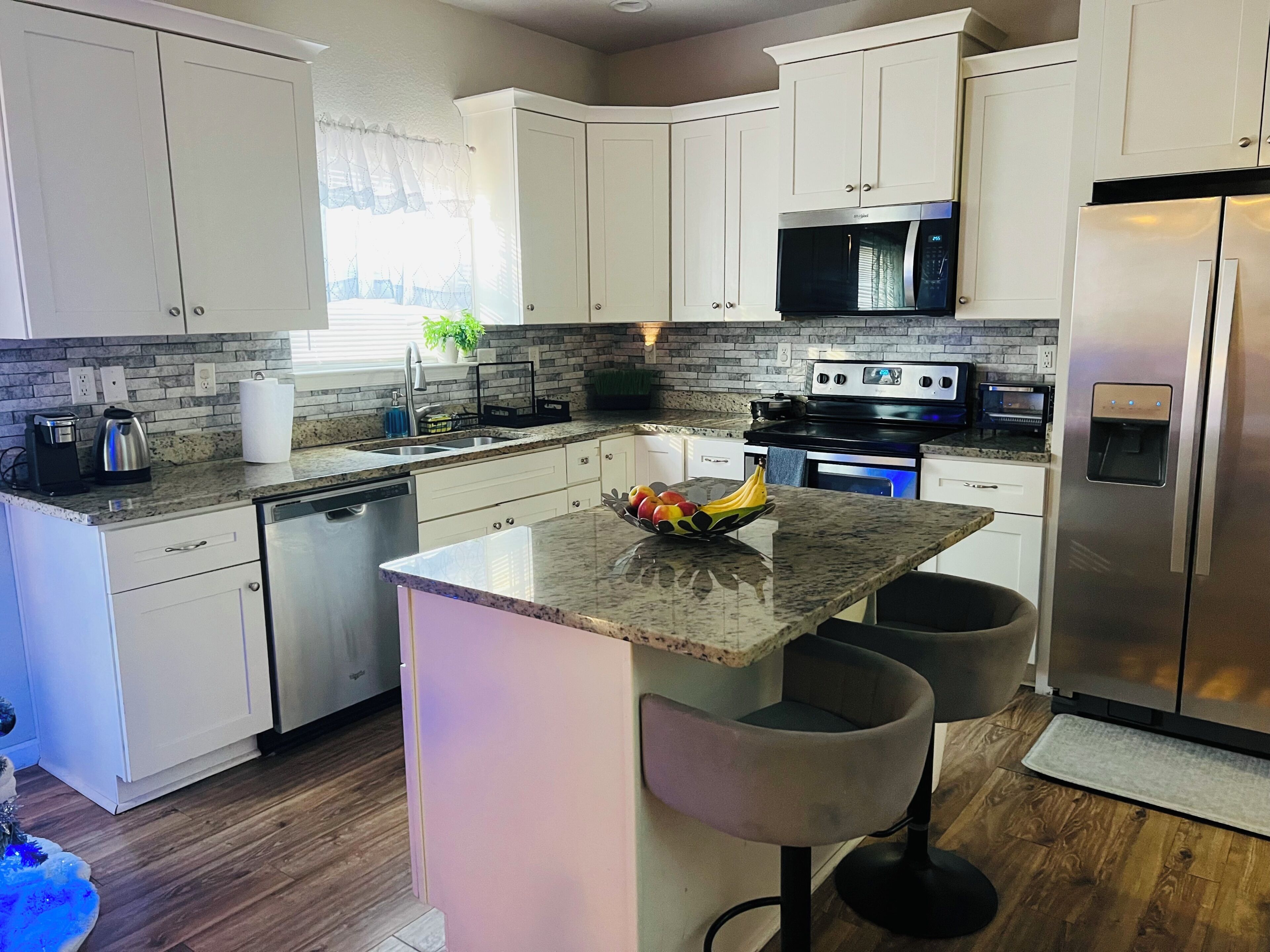 Uma cozinha moderna com armários brancos, bancada de granito, eletrodomésticos de aço inoxidável e uma ilha central com banquetas.