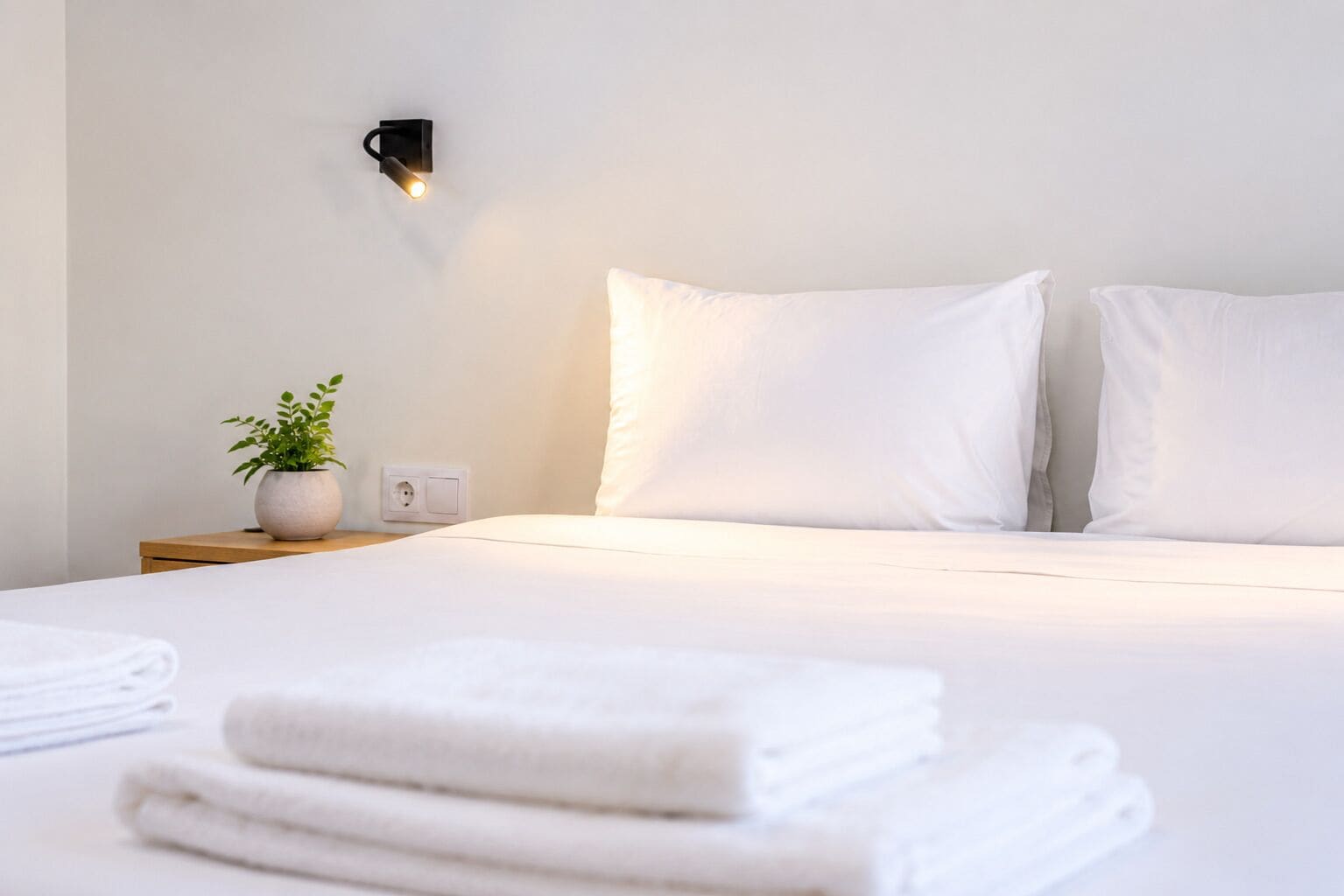 Uma cama bem arrumada com lençóis brancos, uma planta na mesa de cabeceira e um aplique na parede.
