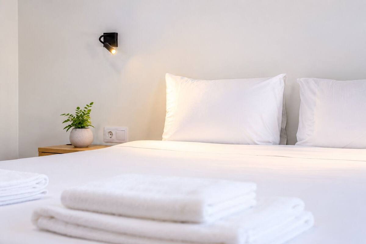 Una cama bien hecha con ropa de cama blanca, una planta en la mesita de noche y un aplique de pared.