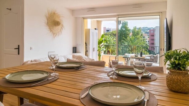 Ein Essbereich mit Holztisch, gedeckt für vier Personen, ein Balkon mit Blick auf Gebäude und Grünflächen sowie ein Fernseher an der Wand.