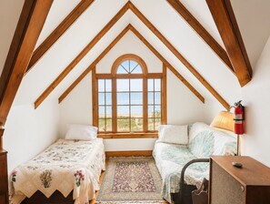 A bedroom with a sloped ceiling, wooden beams, a large window with a view of the beach, two beds, a rug, and a fire extinguisher.