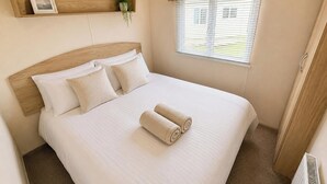 A neatly made bed with two pillows and two rolled towels, a wooden headboard, a small shelf with a picture and a plant, and a window with blinds.