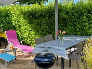 Eine Terrasse im Freien mit einem Tisch, Stühlen, einem Grill und einem Hecken im Hintergrund.