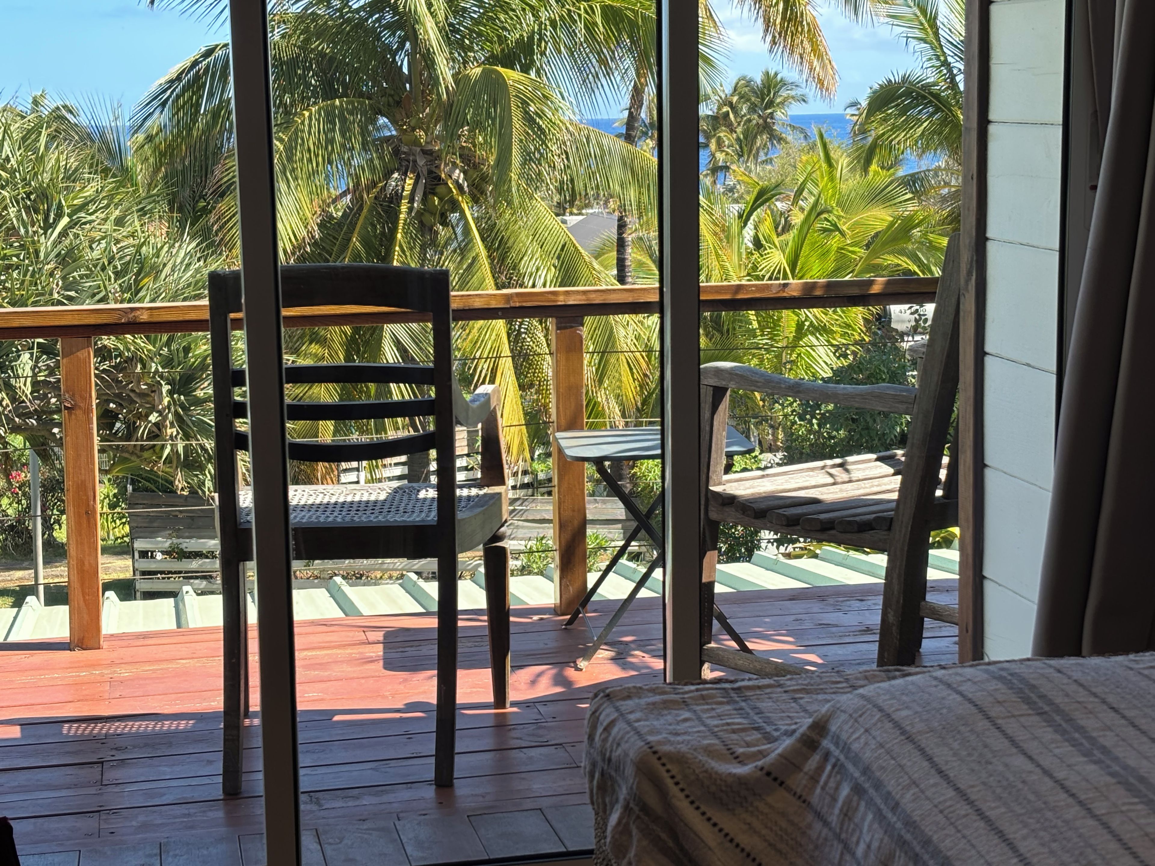 A balcony with two wooden chairs, a wooden table, and a view of palm trees and the ocean.