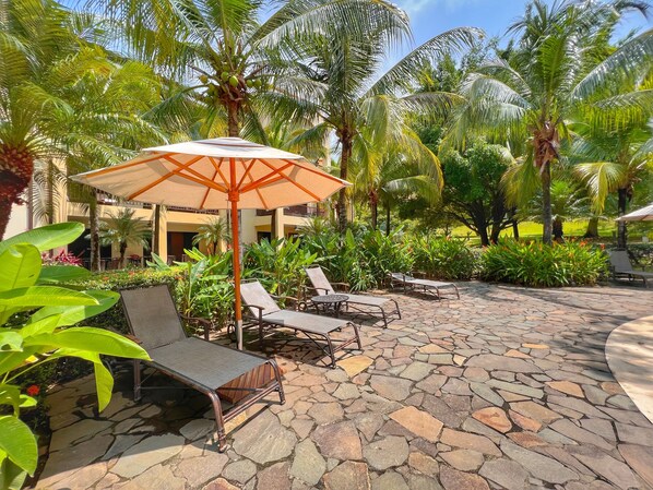 A patio area with lounge chairs and a large umbrella, surrounded by palm trees and lush greenery.