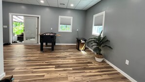 Una habitación amplia con piso de madera, mesa de futbolín, planta en maceta y ventana con persianas.