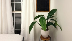 Una habitación con una cama, una ventana con cortinas blancas, una planta en maceta sobre un taburete de madera y una obra de arte enmarcada en la pared.