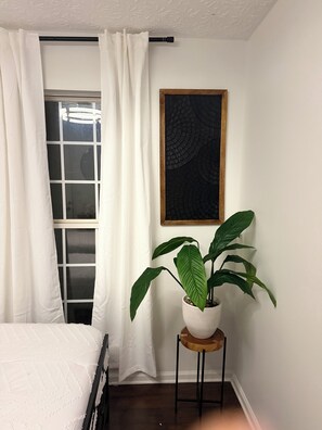 A room with a bed, a window with white curtains, a potted plant on a wooden stool, and a framed artwork on the wall. - Perfect 3-bedroom townhome with AC in charming Decatur (Decatur)