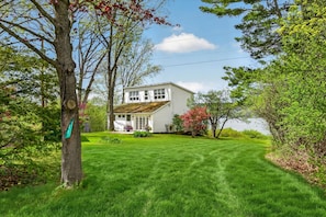 A white house with a well-maintained lawn and a tree with a green sign. - Lakefront retreat with stunning sunsets, scenic lake and mountain view’s (Shoreham)