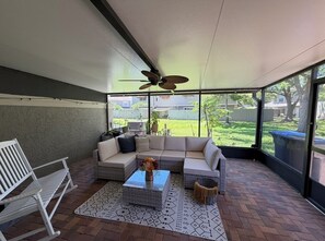 Ein überdachter Patio-Bereich mit einem weißen Sofa, einer weißen Bank, einem gemusterten Teppich und einem Deckenventilator.