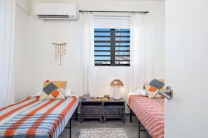 Un dormitorio con dos camas individuales, una ventana con persianas, aire acondicionado instalado en la pared, una mesita con lámpara y un adorno en la pared.