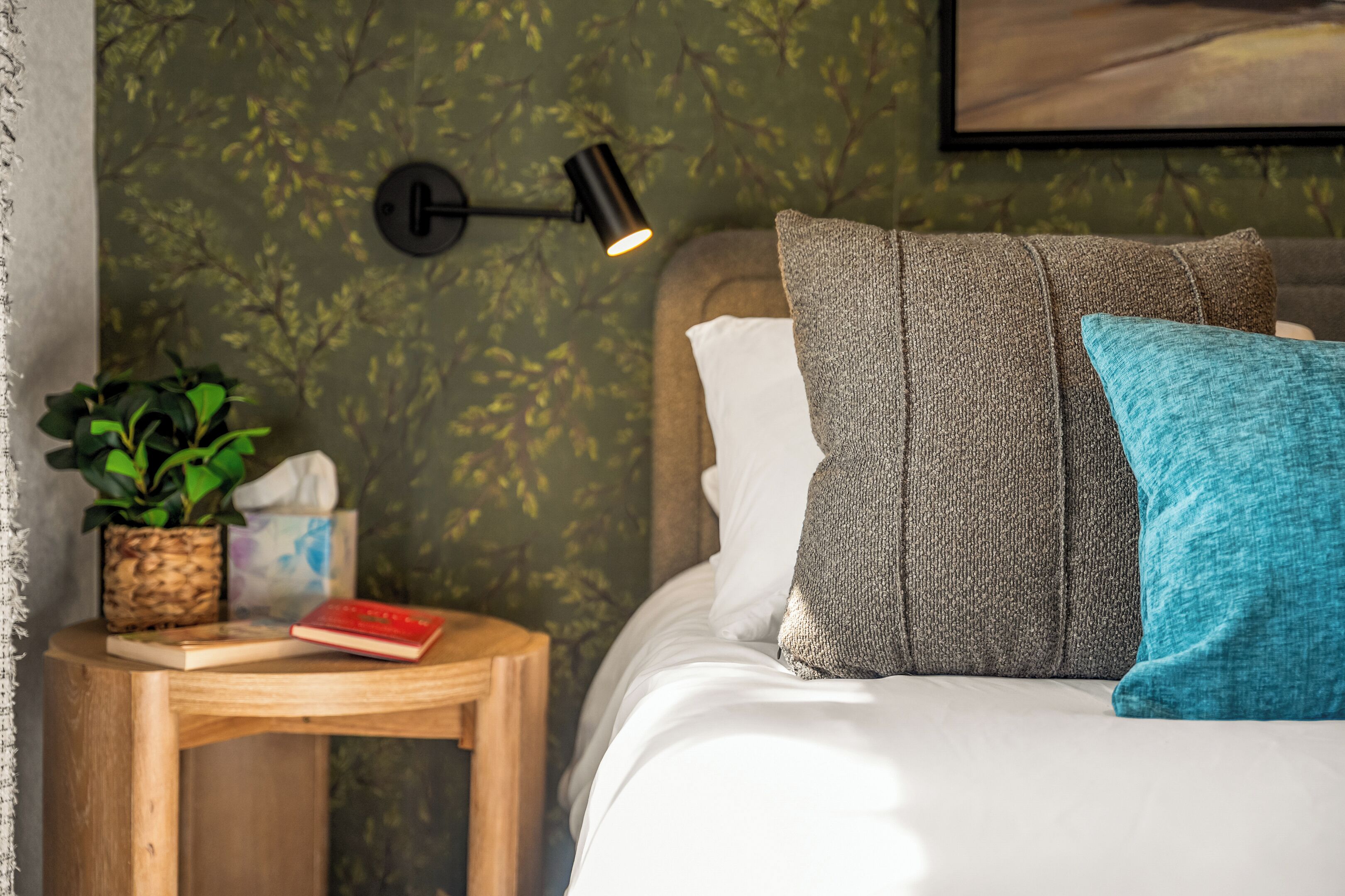 A neatly made bed with pillows, a bedside table with a book and a plant, and a wall-mounted lamp.