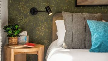 A neatly made bed with pillows, a bedside table with a book and a plant, and a wall-mounted lamp.