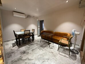 A room with a dining table and chairs, a sofa, a standing fan, and an air conditioning unit.