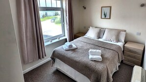 A bedroom with a large bed, a bedside table, a nightstand, a window with curtains, and a wall-mounted picture. - Atlantic Reach Lodge - Spacious 4 Bedroom Retreat (Newquay)