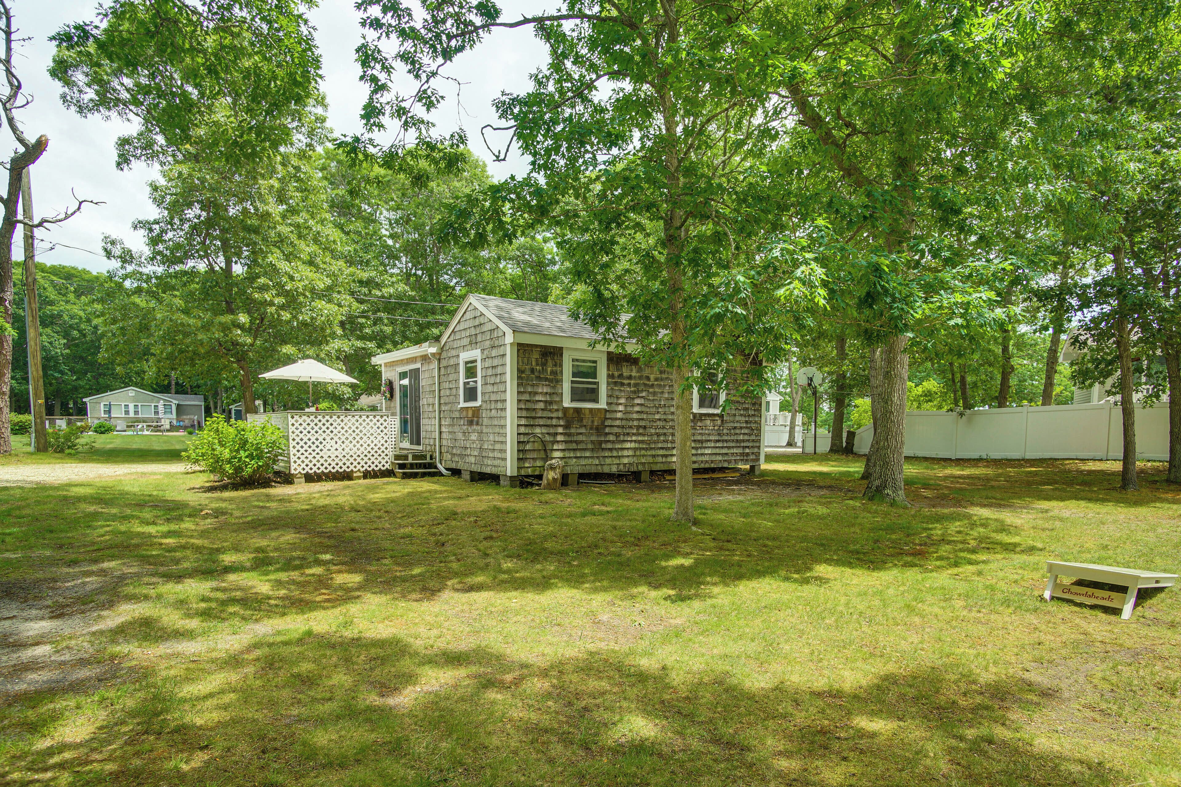 A small wooden cabin with a porch, surrounded by trees and a white fence.