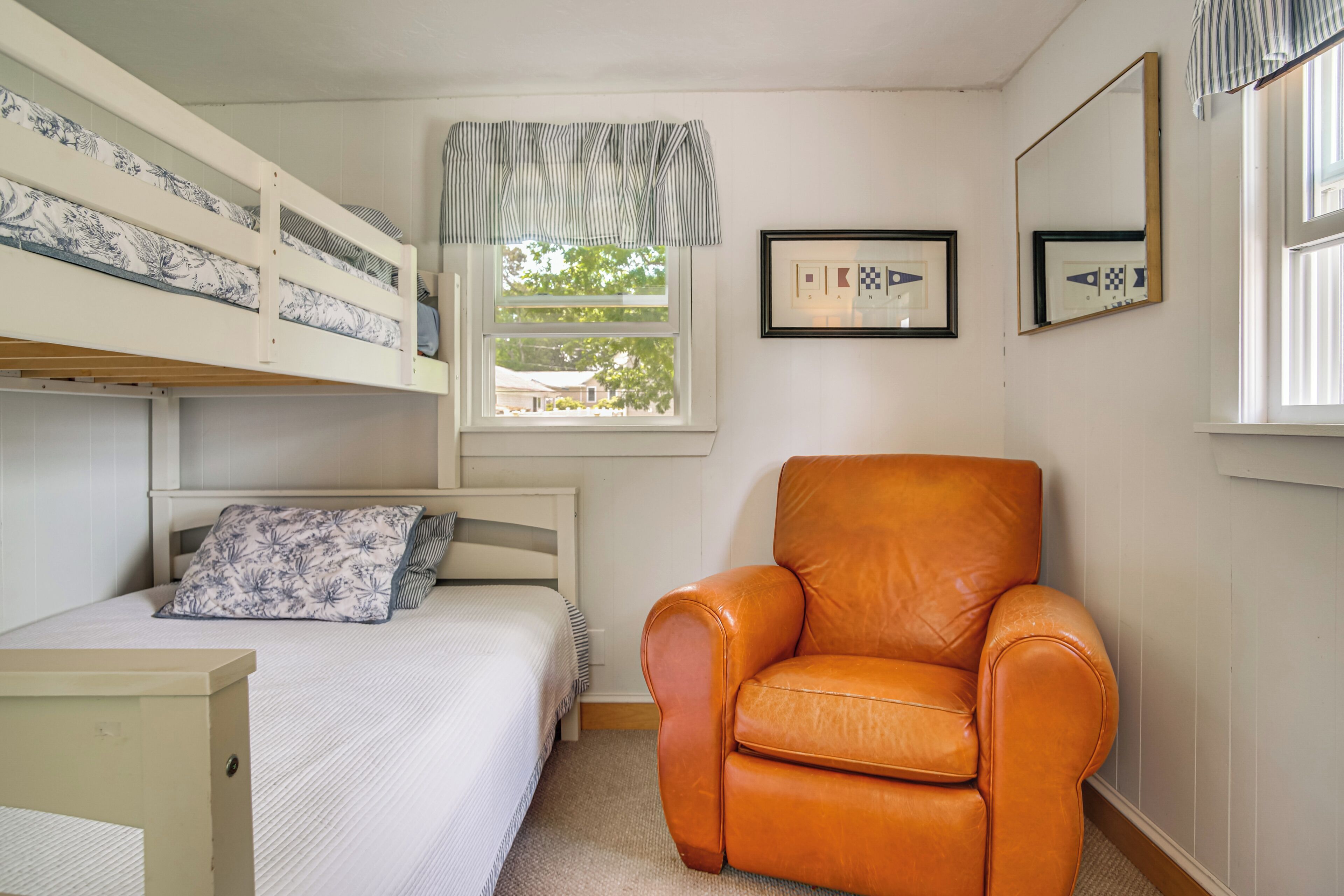 A bedroom with a bunk bed, a single bed, a chair, a window with curtains, and framed pictures on the wall.
