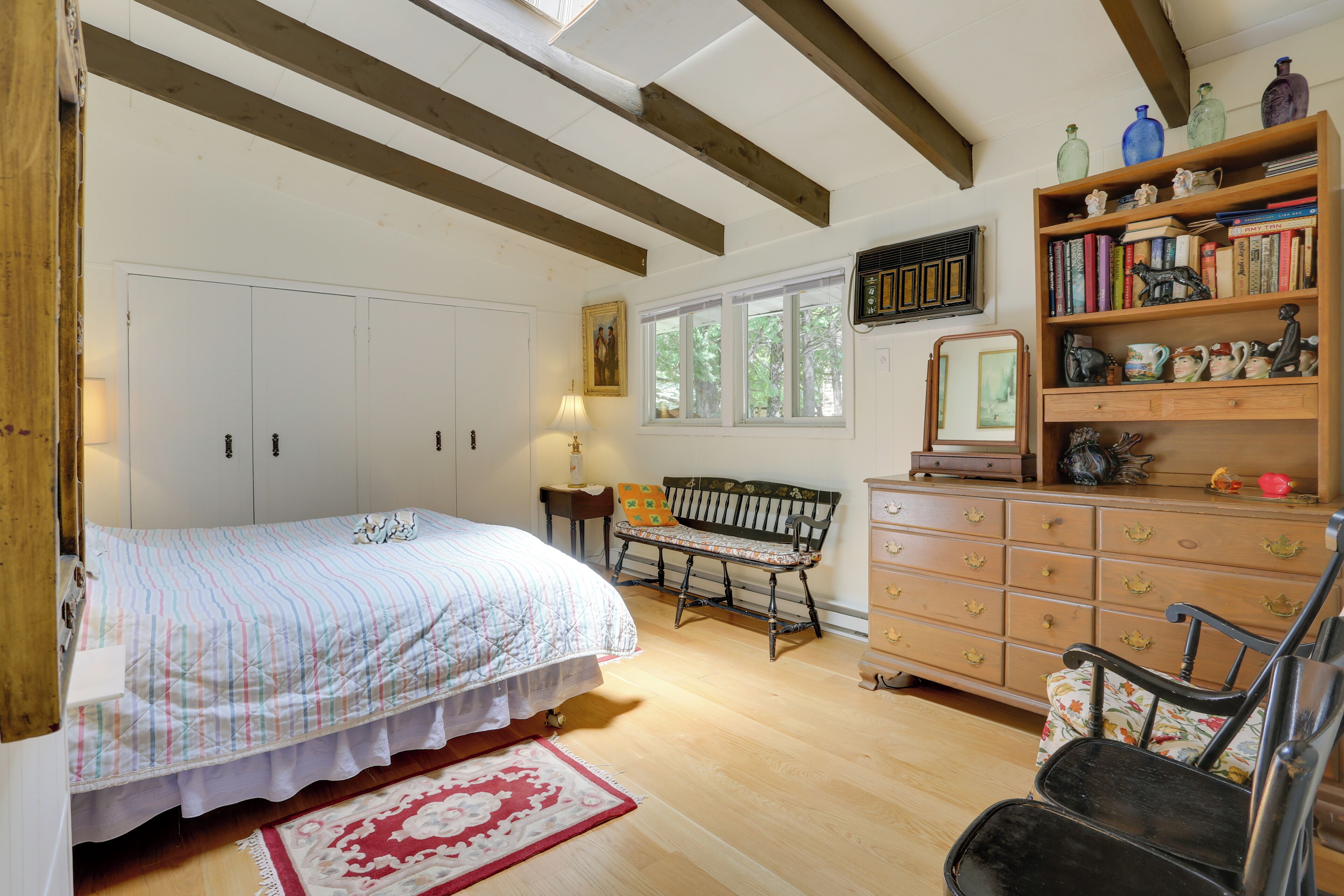 Een slaapkamer met een bed, een houten dressoir, een boekenkast, een bankje en een raam met uitzicht op bomen.