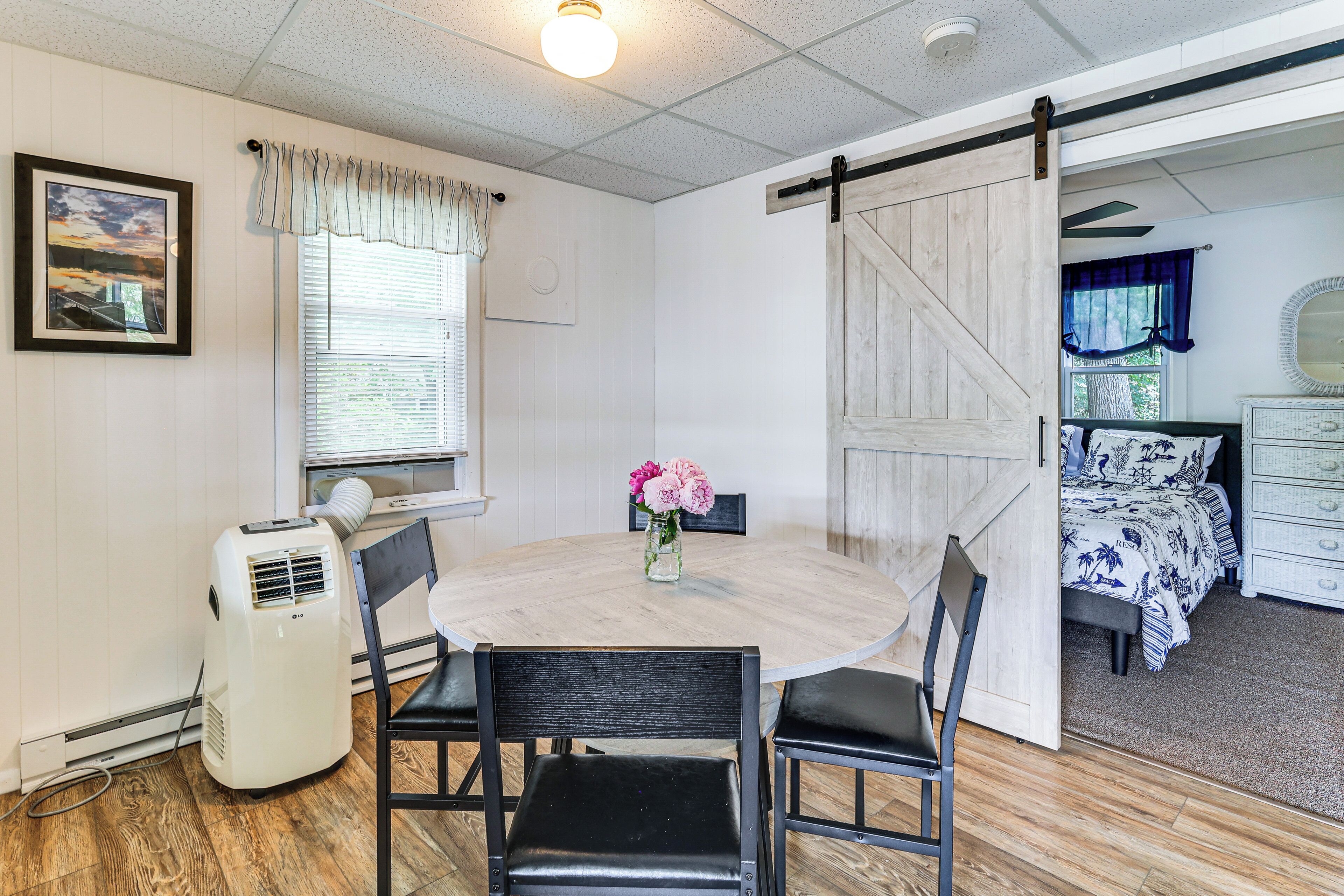 A room with a wooden dining table, four chairs, a white air conditioner, a picture frame, a window with blinds, and a bed with a blue and white floral bedspread.
