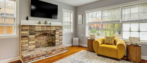 A living room with a brick fireplace, a yellow sofa, a flat-screen TV, and large windows.