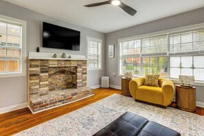 Una sala de estar con chimenea de ladrillo, un sofá amarillo, un televisor de pantalla plana y ventanas amplias.