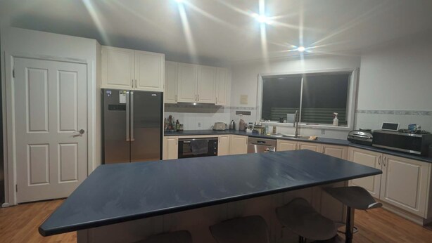 A modern kitchen with white cabinets, a stainless steel refrigerator, a dishwasher, a large black countertop, and a window with blinds.
