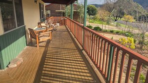 A covered outdoor area with a wooden deck, a seating arrangement, and a view of a landscaped garden and hills.