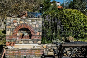 A stone-built outdoor oven with a brick arch, surrounded by greenery and a metal fence.