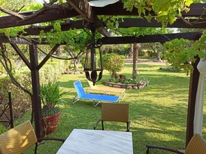 A covered outdoor seating area with a table and chairs, surrounded by greenery and a pergola.