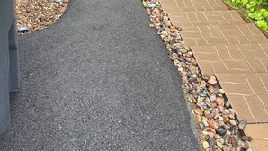 Un sendero pavimentado con borde de guijarros, bordeado por una casa y abundante vegetación.