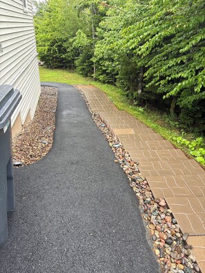 Un sendero pavimentado con borde de guijarros, bordeado por una casa y abundante vegetación.