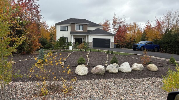 Una casa de dos plantas con garaje, rodeada de follaje otoñal y un jardín ajardinado con rocas y arbustos.