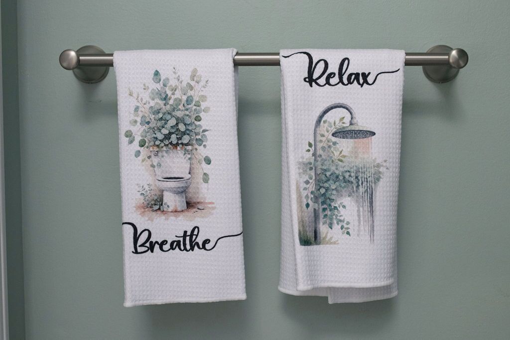 Zwei weiße Handtücher mit floralen Mustern und den Aufschriften 'Breathe' und 'Relax' hängen an einem Metallhandtuchhalter.