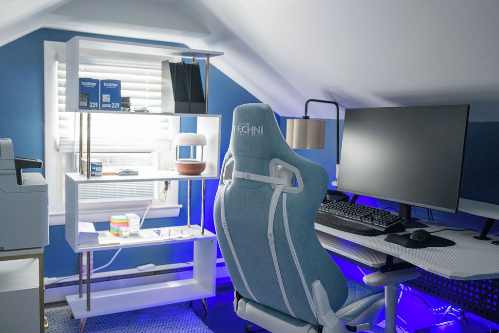 Ein moderner Gaming-Stuhl mit sichtbarem Markennamen, ein Schreibtisch mit Computer-Einrichtung und ein Regal mit Gegenständen, darunter eine Kiste und eine Pflanze.