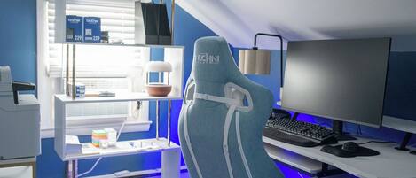 A modern gaming chair with a visible brand name, a desk with a computer setup, and a shelf with items including a box and a plant.